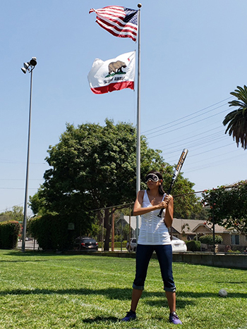 A female batter is blindfolded and in batting stance with the American and California flags visible in the background.