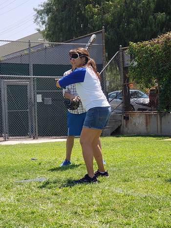 A female wearing a white shirt with blue sleeves stands in batting stance.