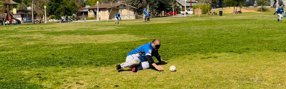 Bee Yang is on his left hip on the ground, reaching out for a beep baseball just in front of him. He is wearing gray baseball pants, a blue Panthers jersey, and a blindfold. He has a very short buzz cut. Bee is on a large grass field in a park. Way back in the distance there are several other panthers players, Brendan Wright, Joe Duncan, and Leo Medina. Spotter Jeff Collins stands between the players in right field. The edge of the field is lined with several small trees, light posts, and a small bathroom building.